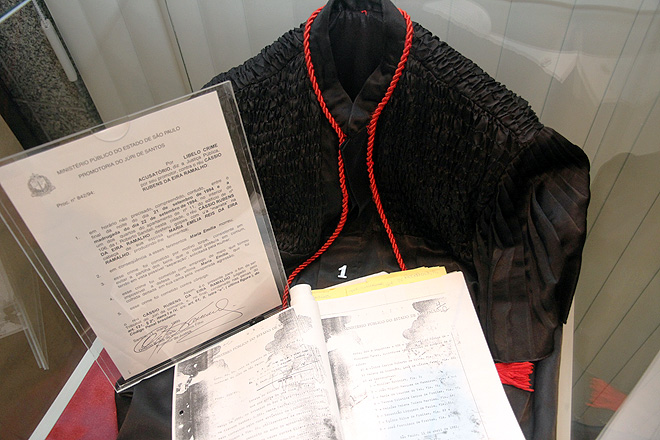 uma roupa preta de tribunal com detalhe vermelho, uma placa em acrílico com um documento dentro contextualizando (ilegível) e um processo aberto em cima (ilegível)