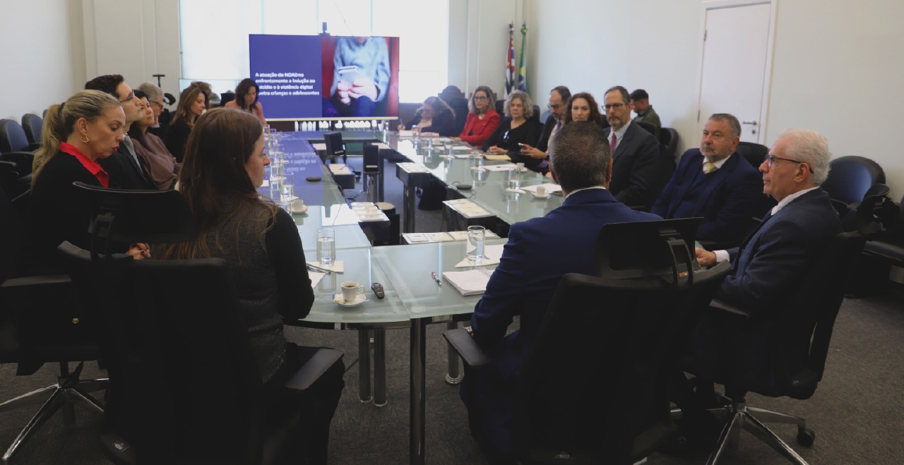 Autoridades presentes no evento reunidas em uma mesa em frente a telão